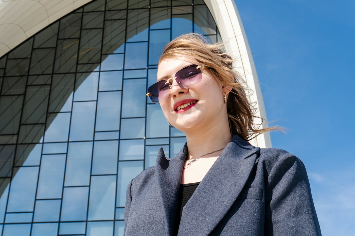 Woman in a blazer and sunglasses stands before a modern building.