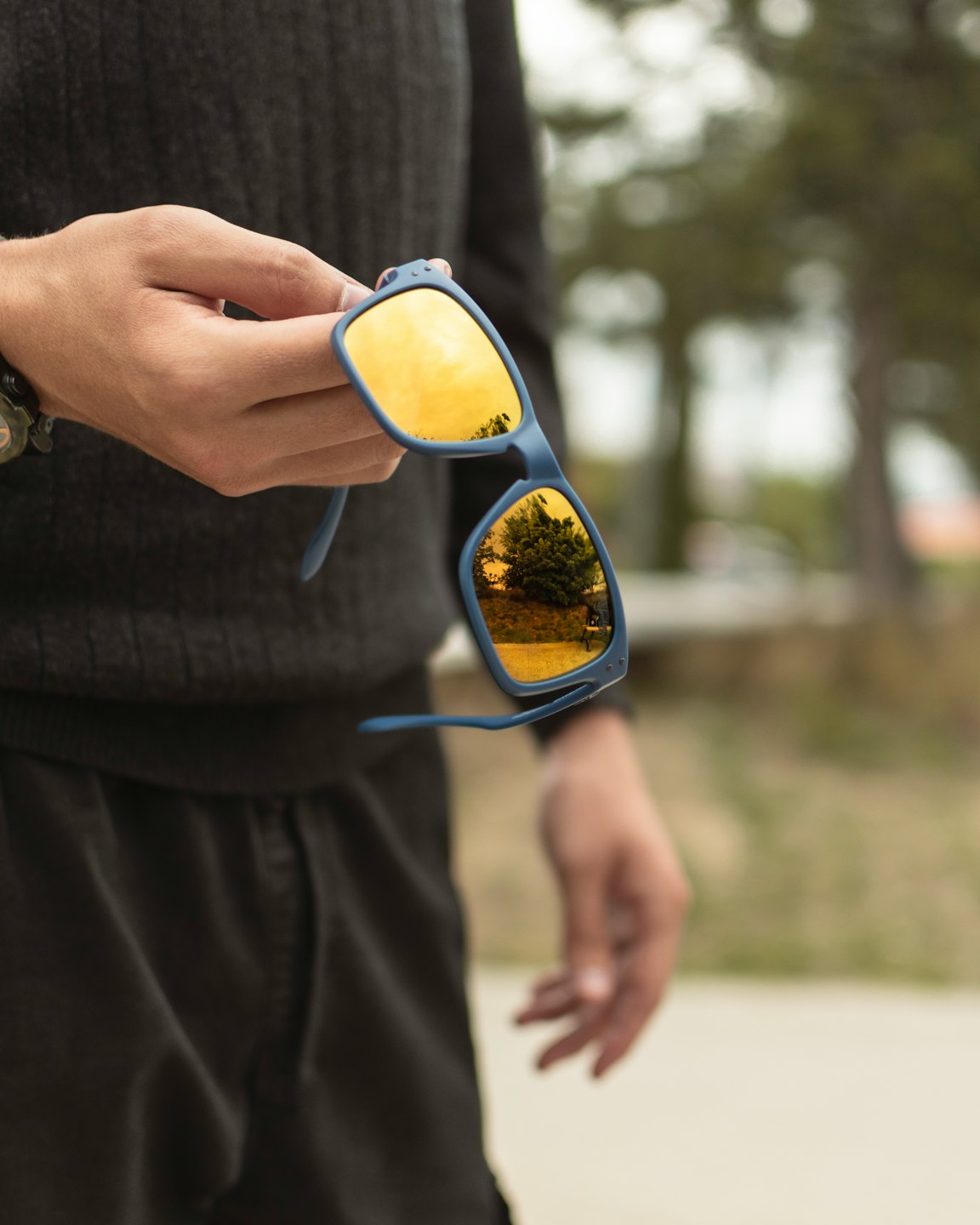 A hand holds blue sunglasses reflecting trees and a field.
